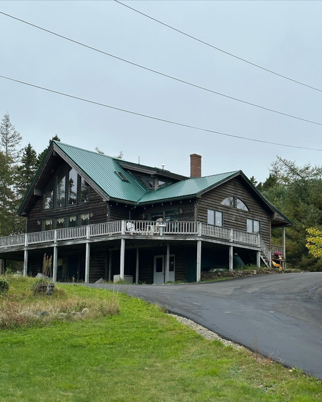 Log cabin with green roof, wraparound deck, and chimney under cloudy sky.