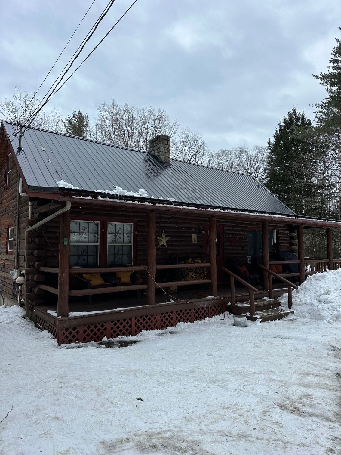Log cabin with metal roof, snow on the porch and ground, overcast sky.