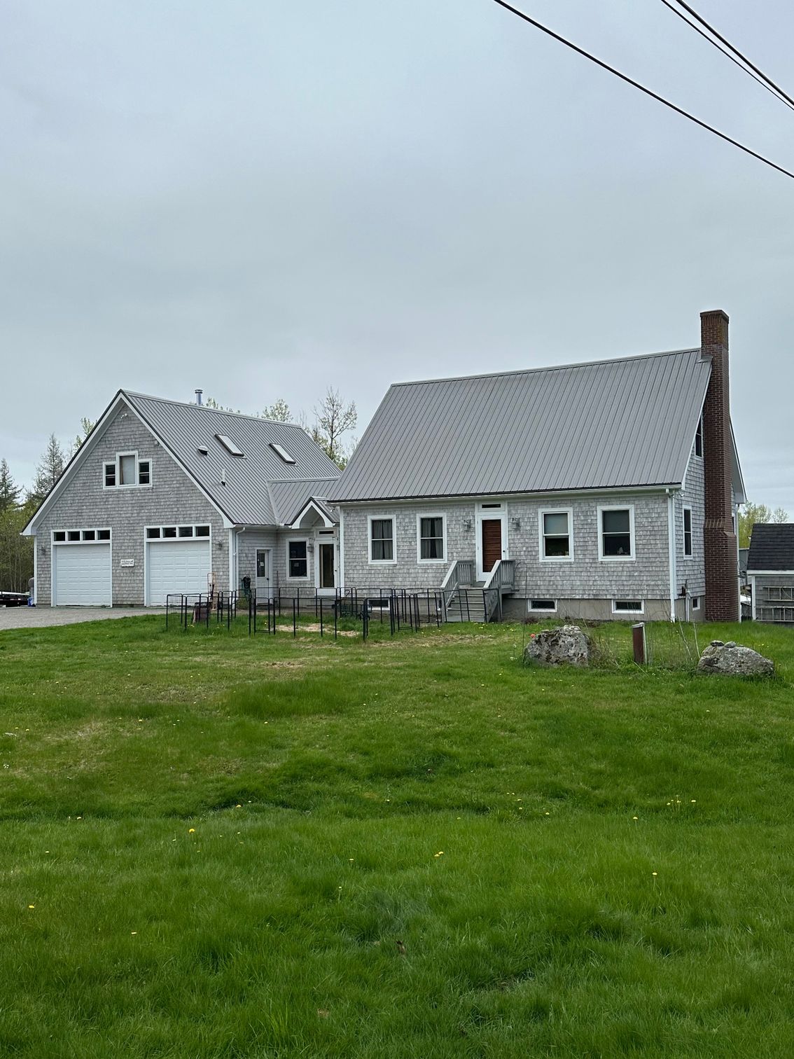 Gray shingled house with garage and chimney on a grassy lot under an overcast sky.