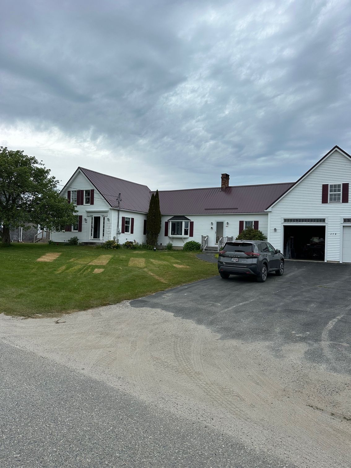 White farmhouse with red roof and shutters, gray car parked on driveway, cloudy sky.