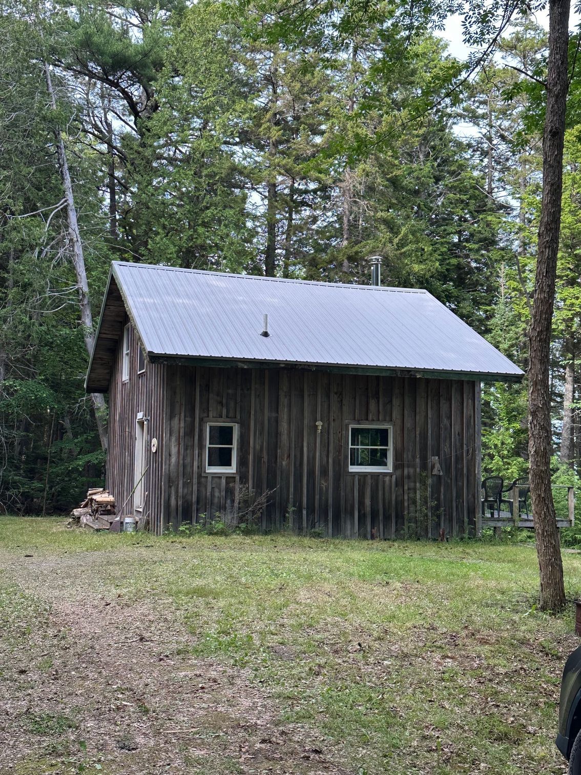 Weathered wooden cabin in a wooded area with two windows, a metal roof, and a chimney.