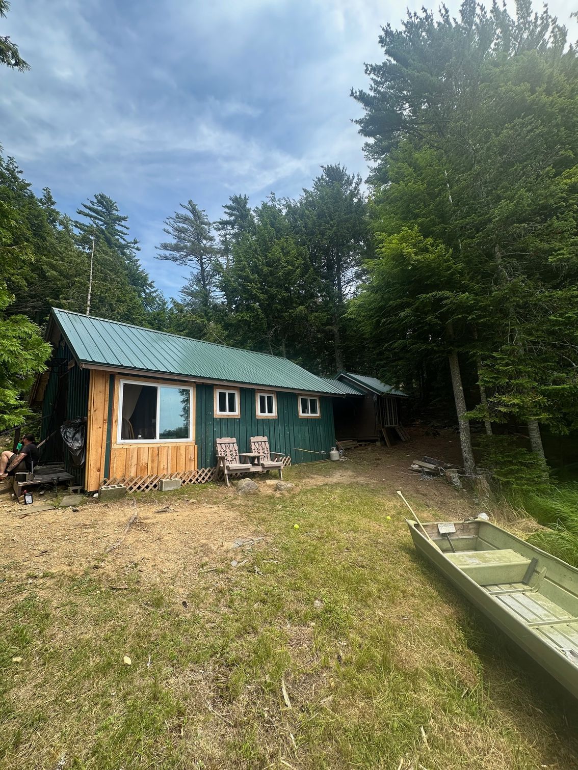 A small green cabin on a grassy shore next to a boat, surrounded by trees. Cloudy sky.