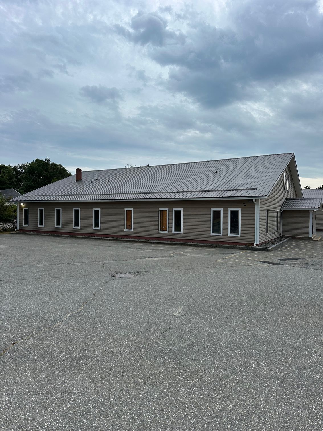 Tan commercial building with a gray roof and numerous windows, set on a gravel lot under an overcast sky.