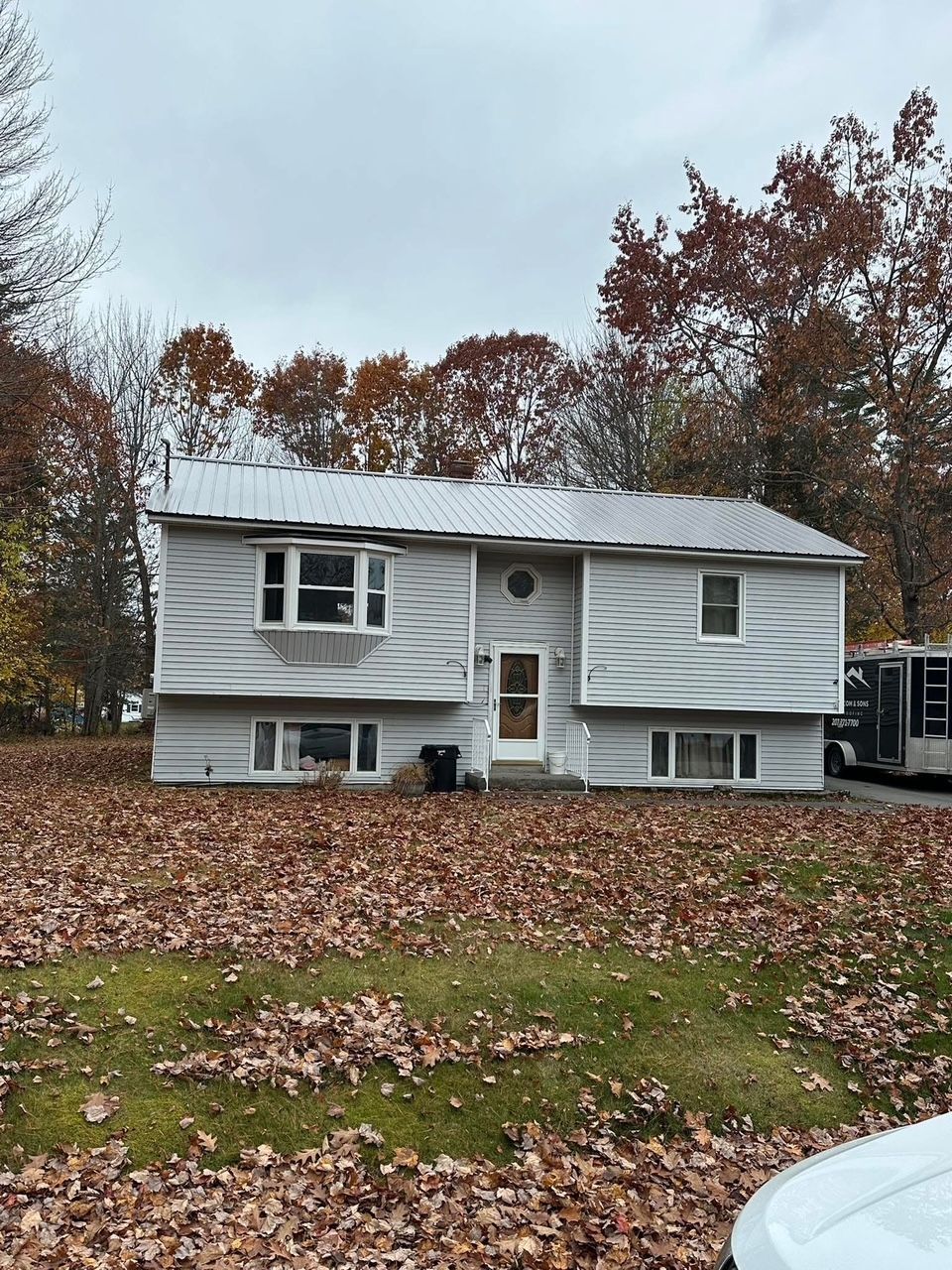 Light gray split-level house with fall foliage on the ground and trees, overcast sky.