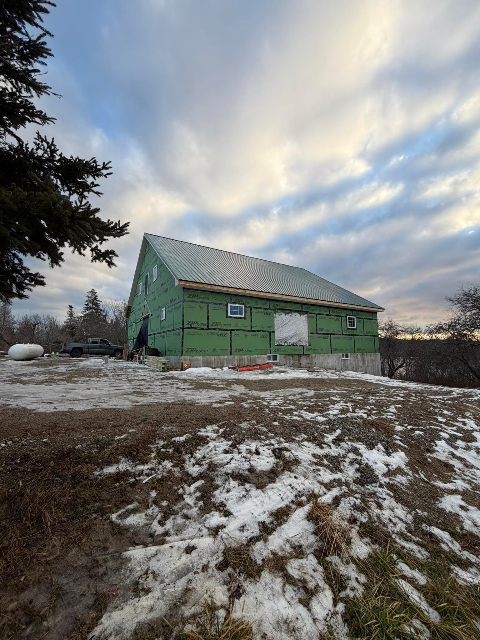 Building under construction with green siding and partial roofing, set in a snowy, overcast environment.
