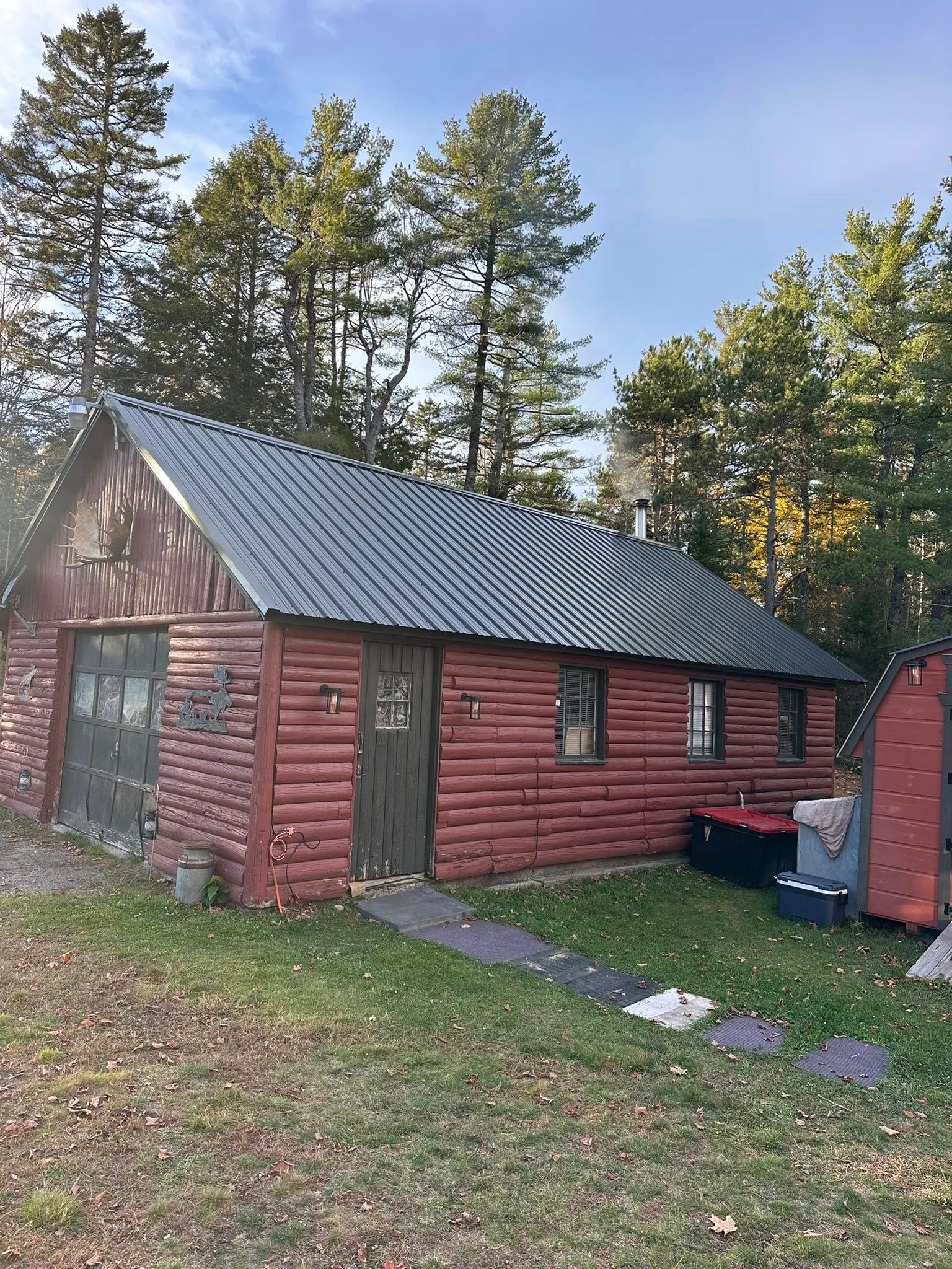 Red log cabin with a corrugated metal roof, a garage door, and trees in the background.