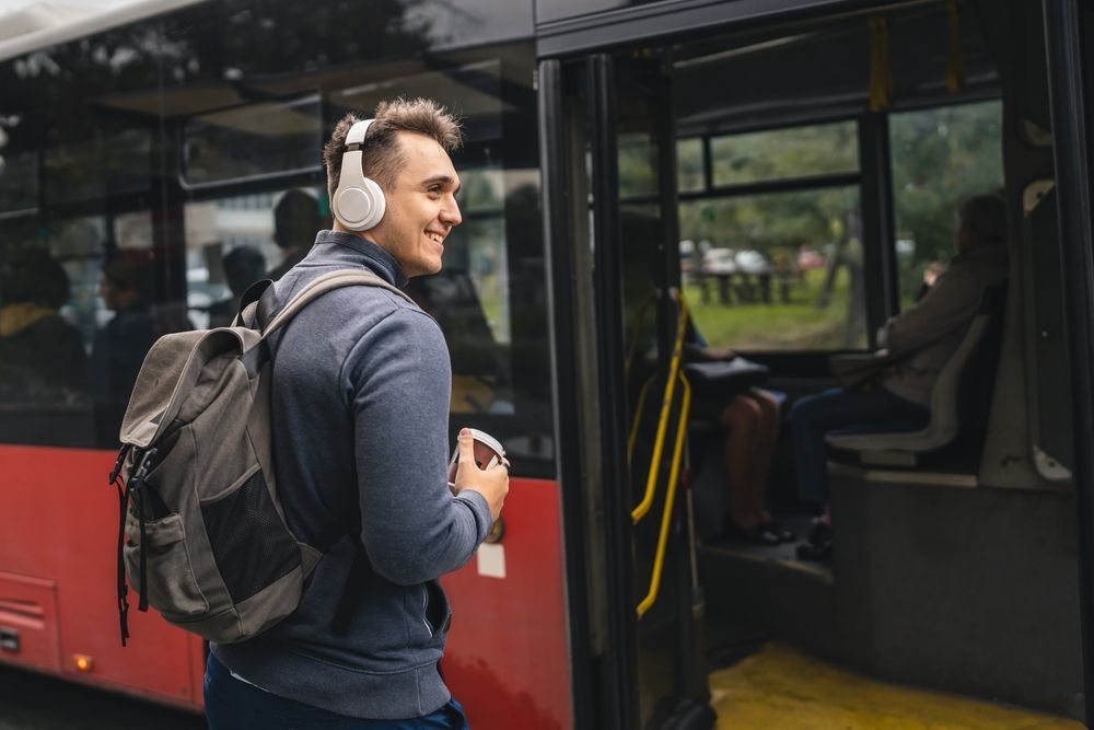 A young adult male is getting on a bus with headphones and a backpack.