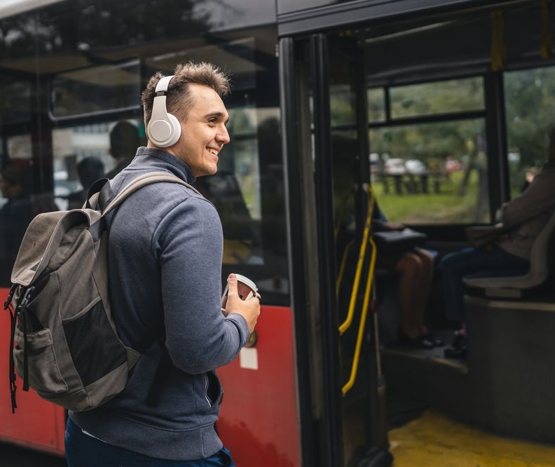 A young adult male is getting on a bus with headphones and a backpack.