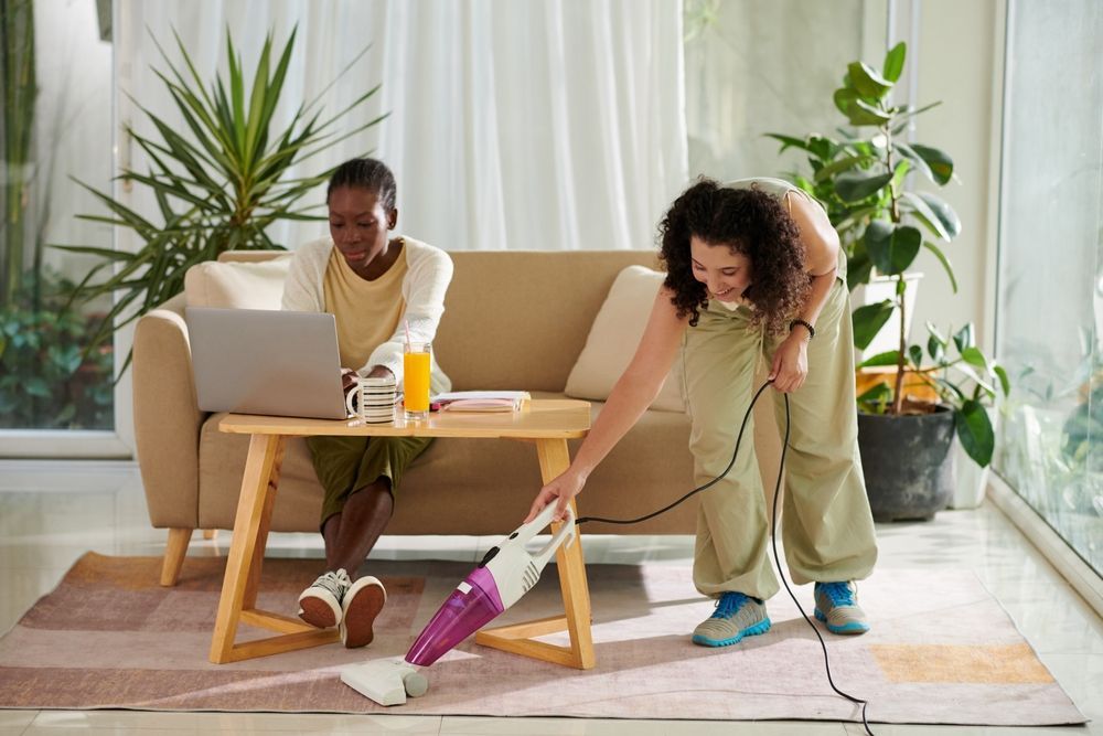 Cheerful girl vacuum cleaning carpet when her roommate studying for exam