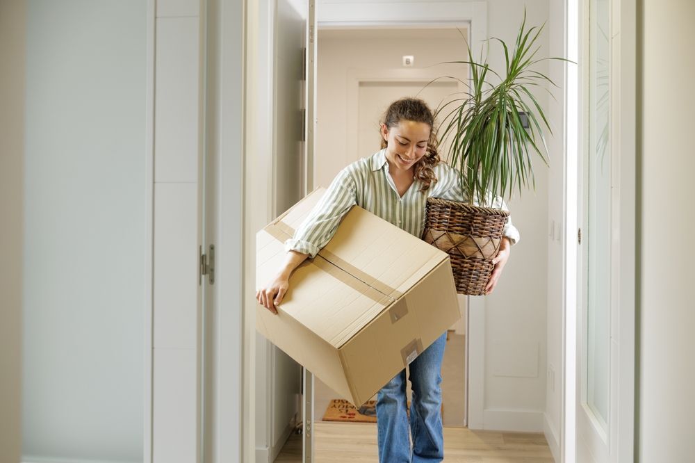 woman carrying moving box and plant through her apartment