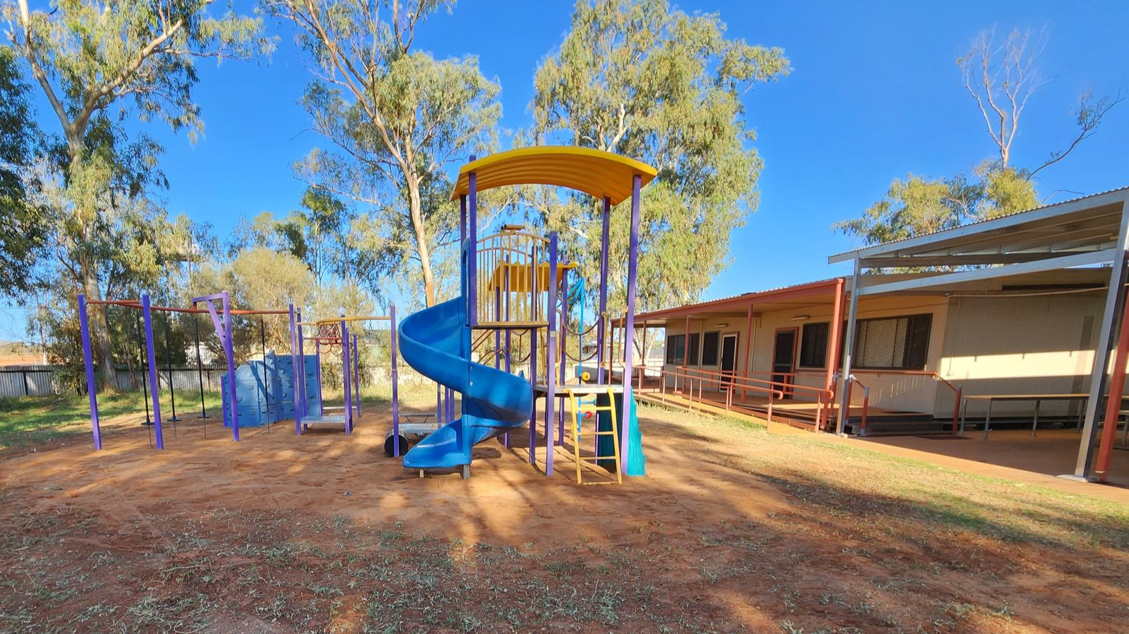 A bright blue spiral slide on a playground with tan ground, surrounded by trees next to a building on a sunny day.