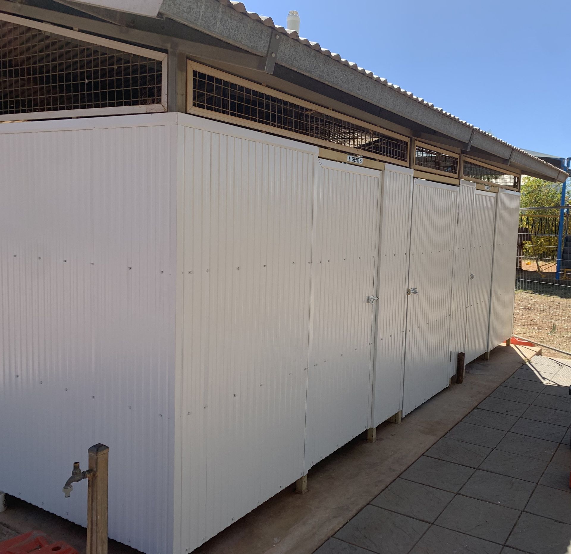 A white corrugated metal structure with multiple doors, possibly public restrooms or stalls, set outdoors.