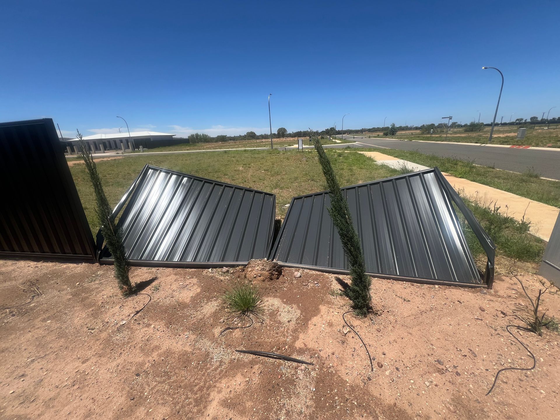A broken, dark-colored corrugated metal fence lies flat on the ground in an empty, sunny, dirt-covered lot.