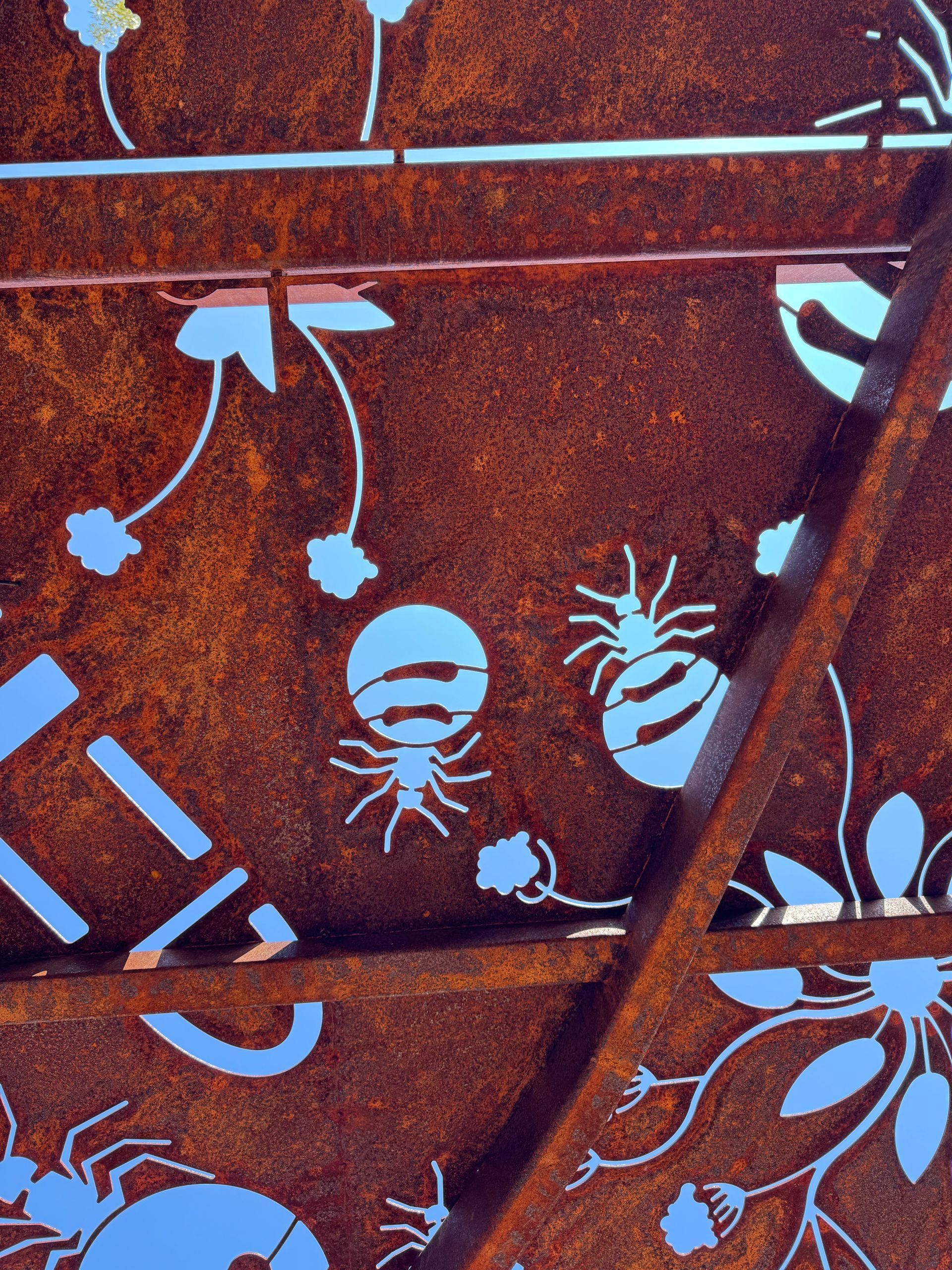 A rusty metal panel features cutout patterns of spiders, flowers, and vines against a light-colored background.