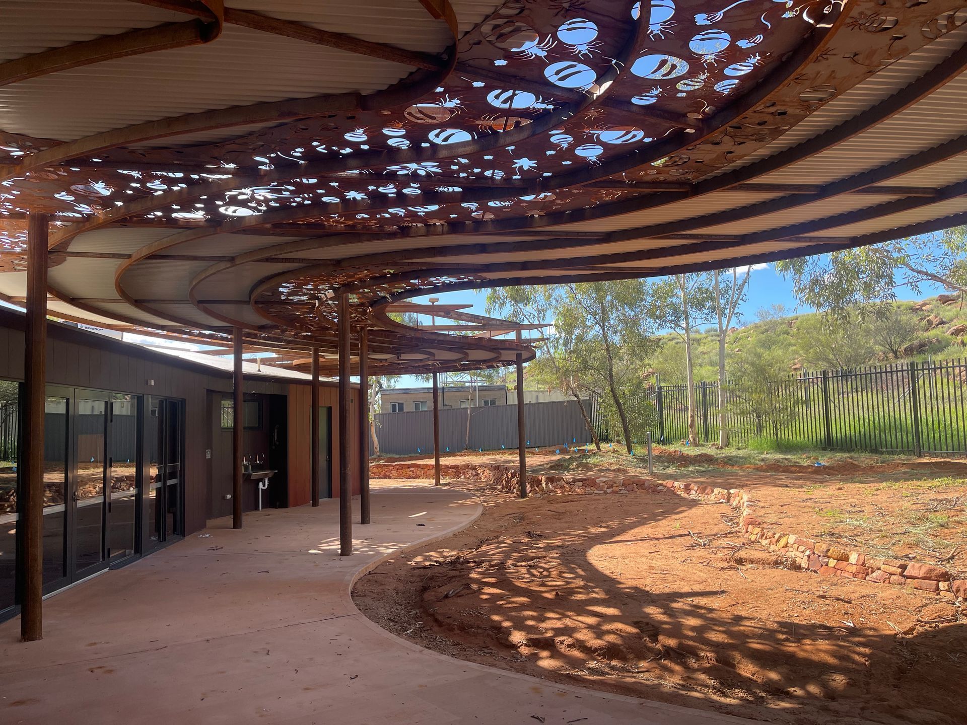 An outdoor patio with a patterned, rusted-metal canopy providing shade over a curved pathway in a desert landscape.