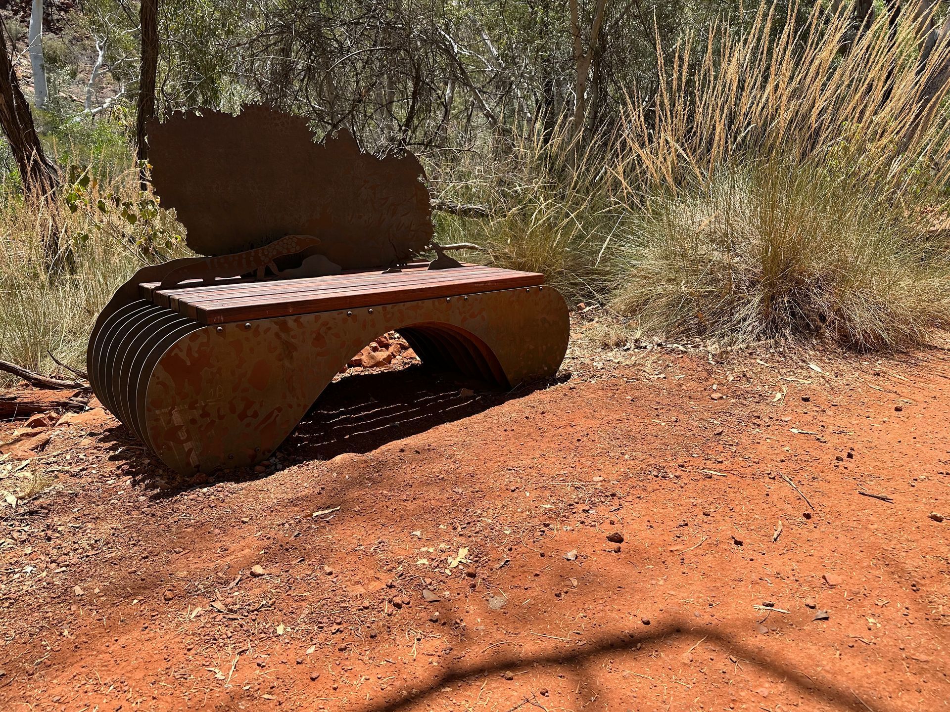 A textured, rust-colored metal bench with an abstract, organic-shaped backrest sits on a red dirt path in a wooded area.