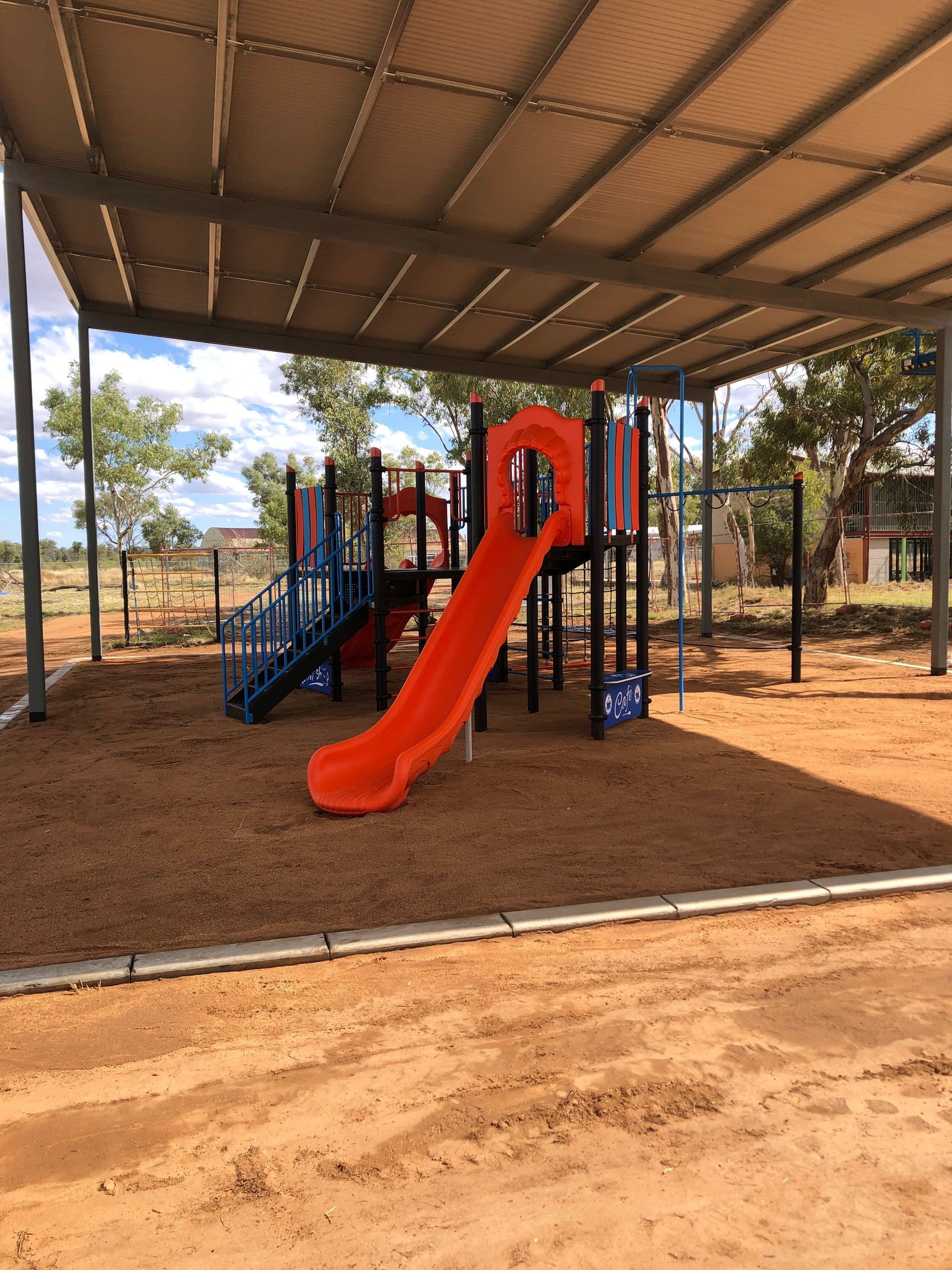 A colorful playground structure with a bright orange slide under a metal shade canopy on a wood-chipped ground.