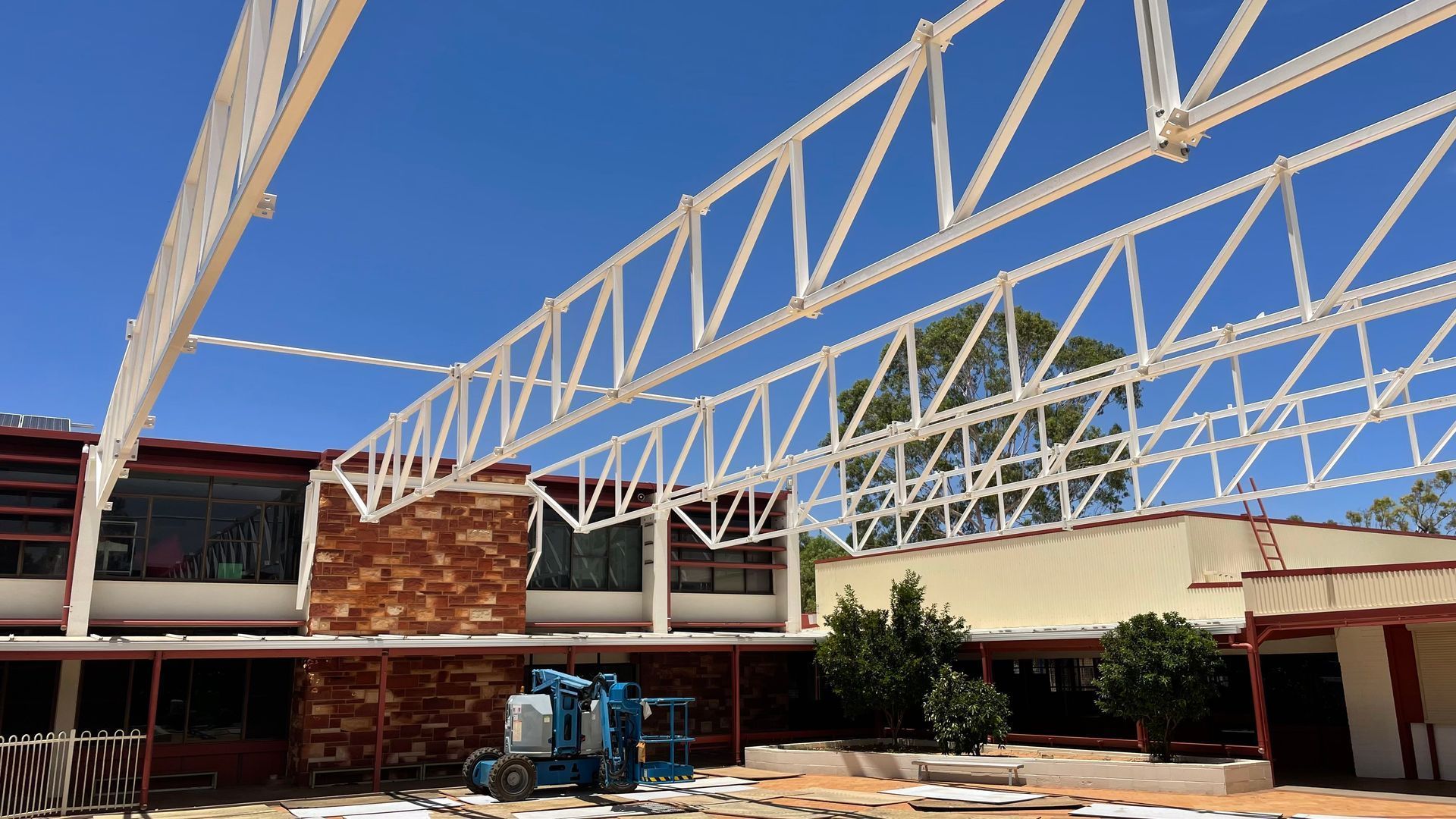 Large white metal trusses span the outdoor courtyard of a building under a bright blue sky.