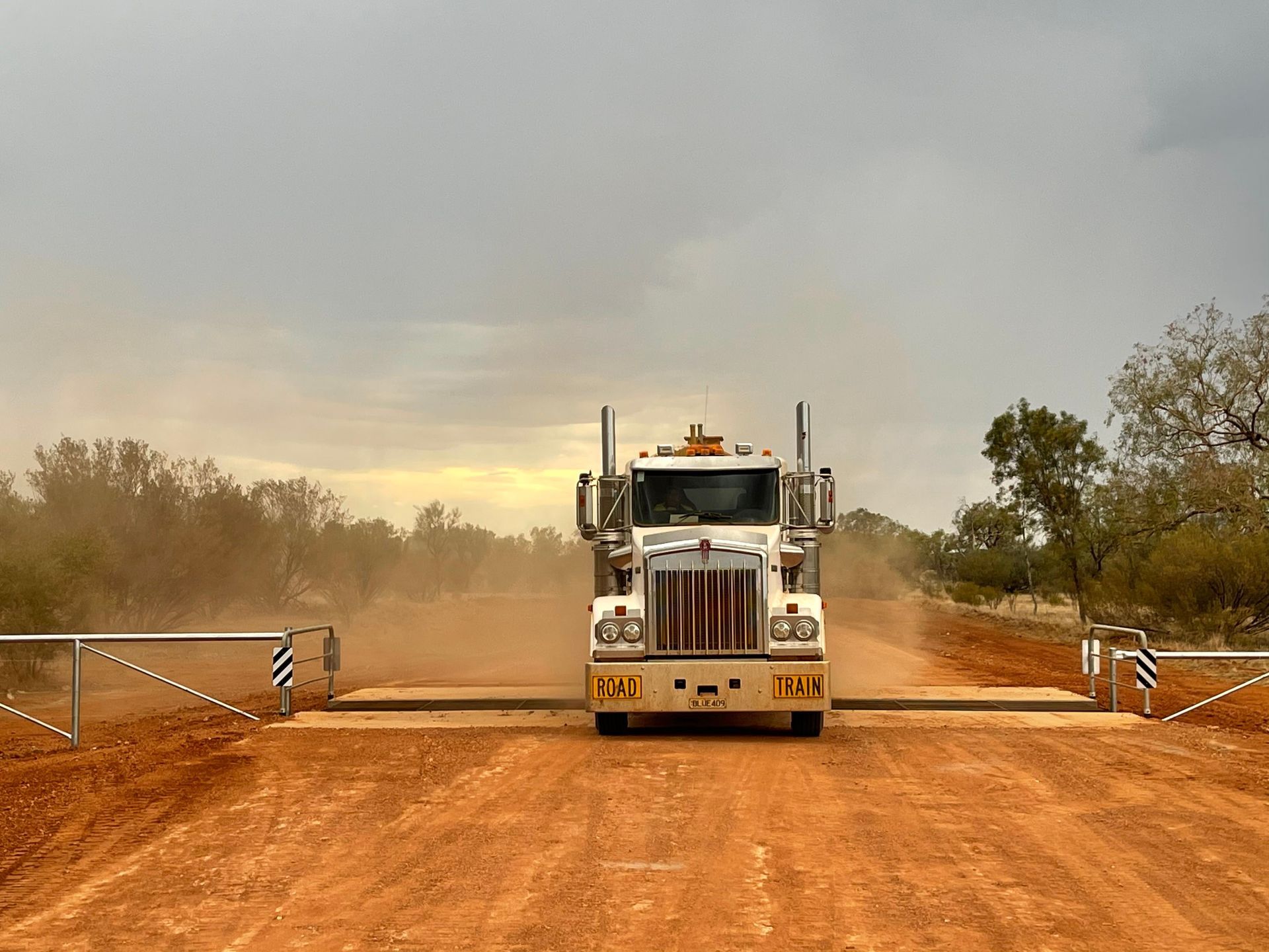 A white semi-truck drives over a cattle guard on a dusty red dirt road under a cloudy sky.