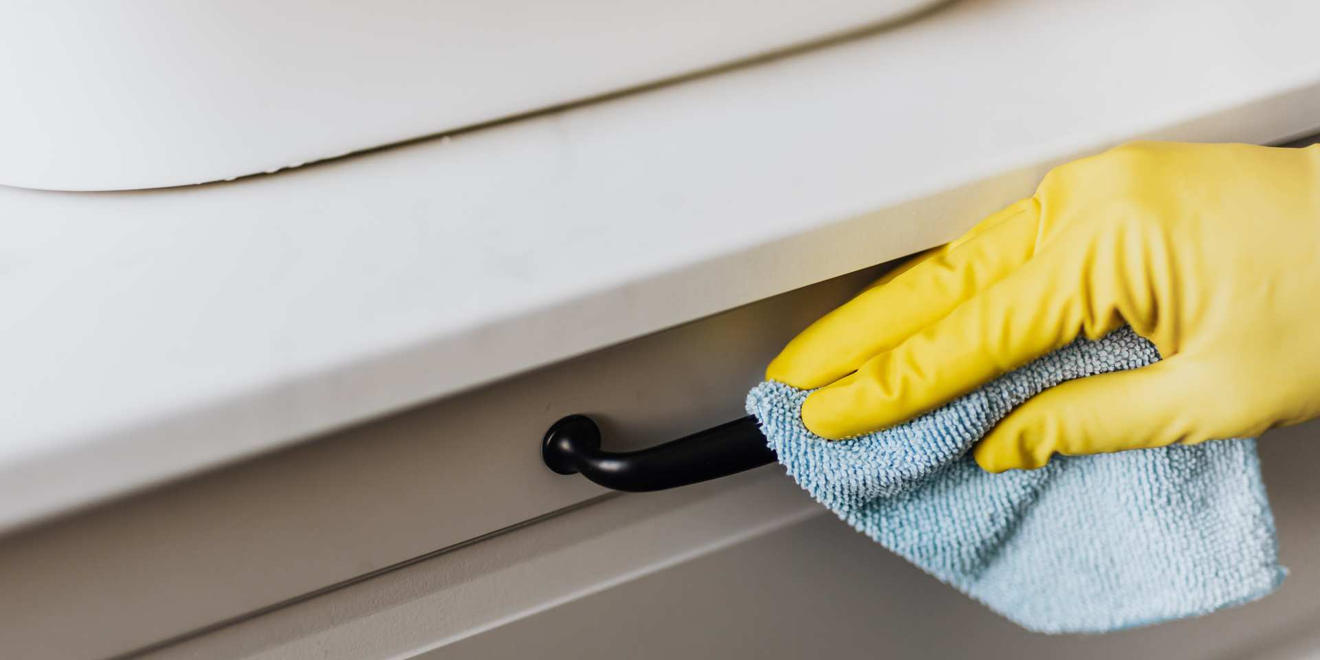 A hand in a yellow glove cleans a cabinet pull with a blue cloth.