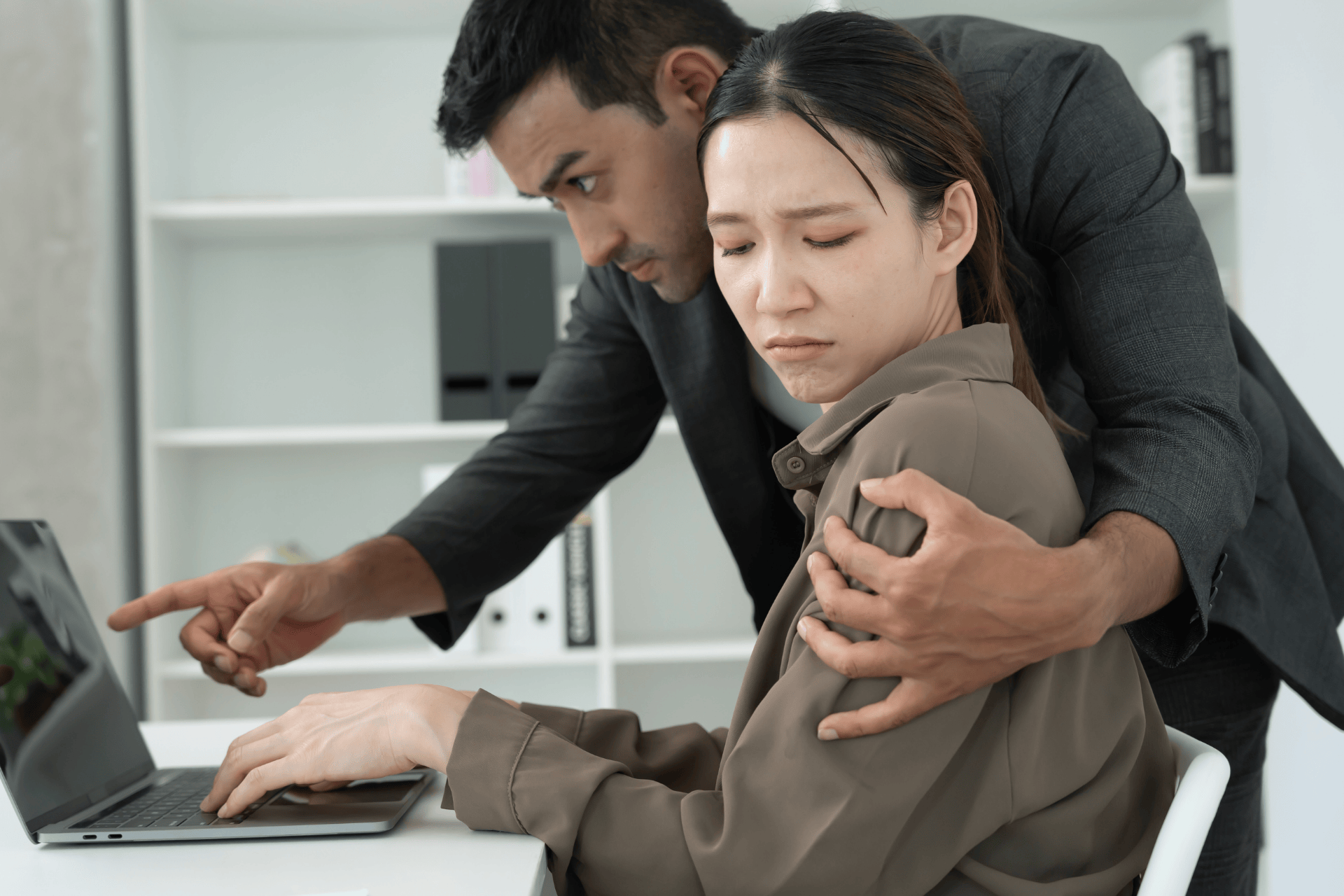 A man stands closely behind a seated woman, holding her shoulder and gesturing toward her laptop as she appears uneasy at her desk in a modern office.