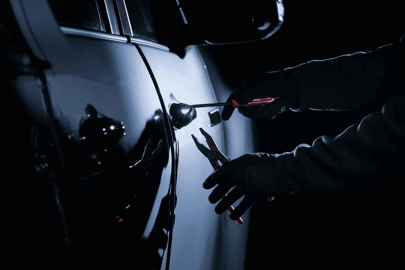 A car thief is using tools to break into a vehicle door at night.