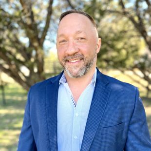 A man in a blue suit and blue shirt is standing in front of a tree.