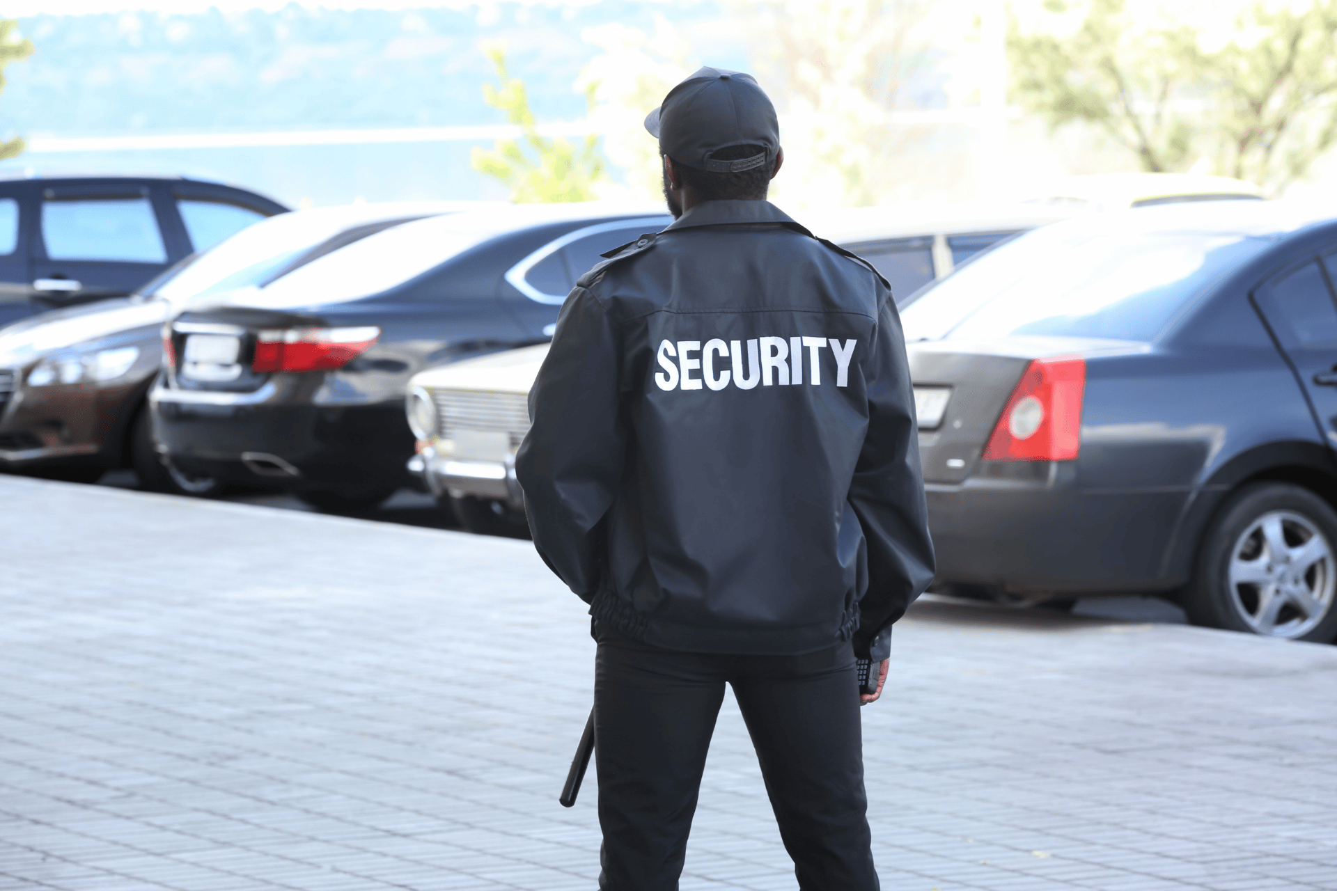Security guard standing in a parking lot monitoring parked vehicles, wearing a black jacket labeled “SECURITY” while conducting patrol duties.
