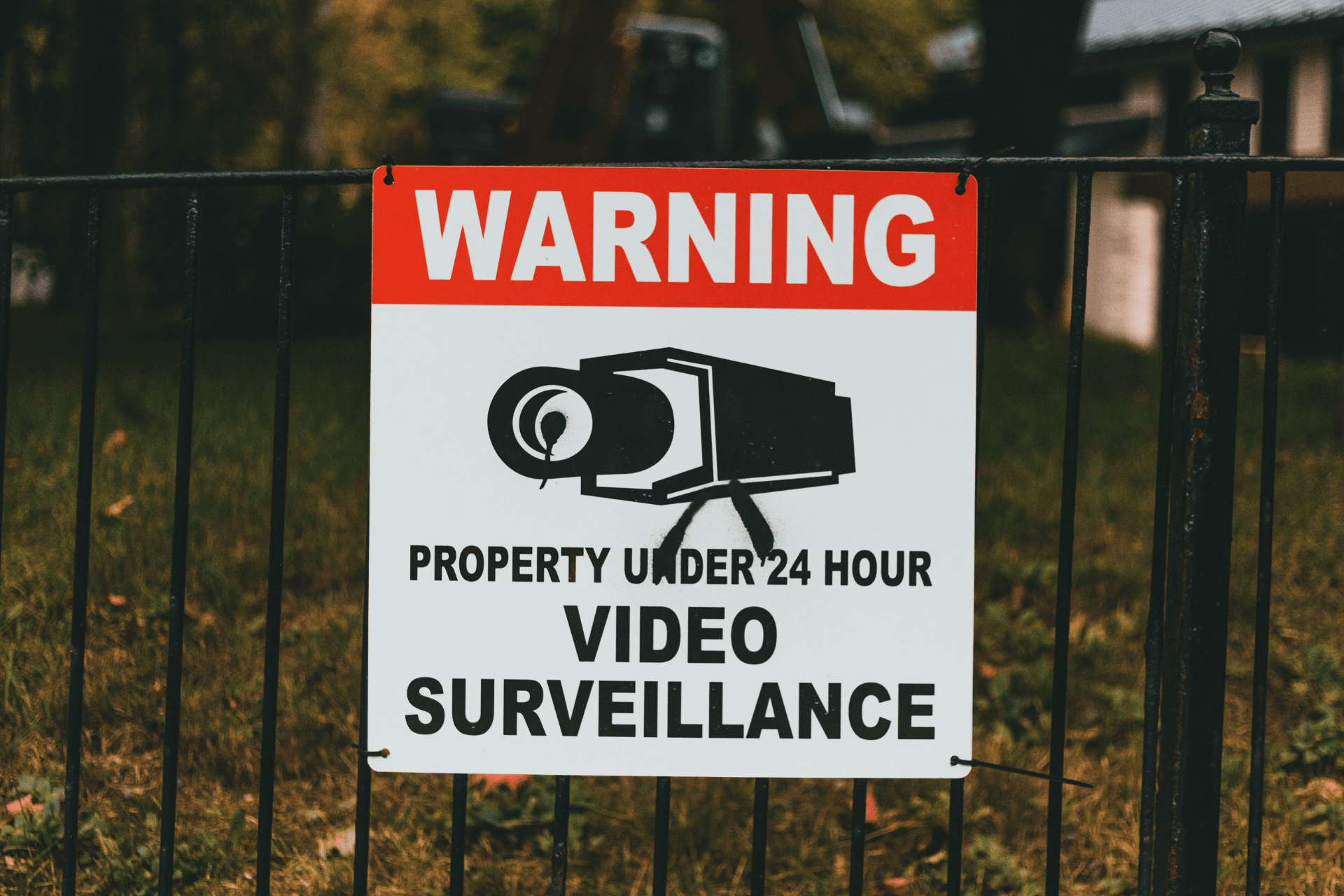 Warning sign on a fence indicating the property is under 24-hour video surveillance with a security camera symbol.