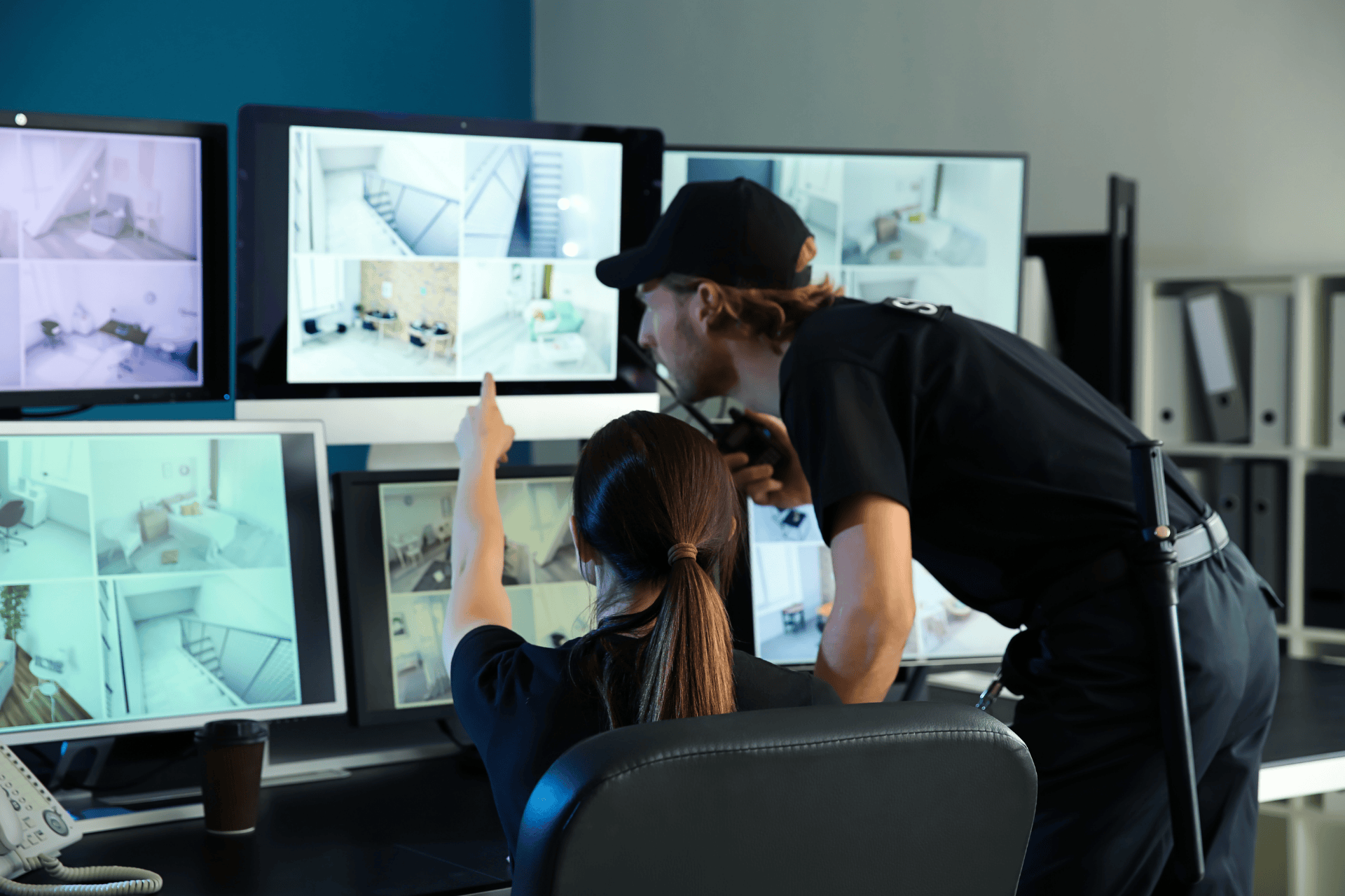 Security personnel are monitoring multiple surveillance camera feeds on computer screens in a control room while coordinating a response.
