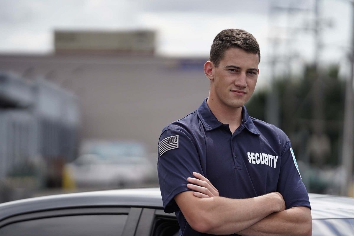 A security guard is standing next to a car with his arms crossed.