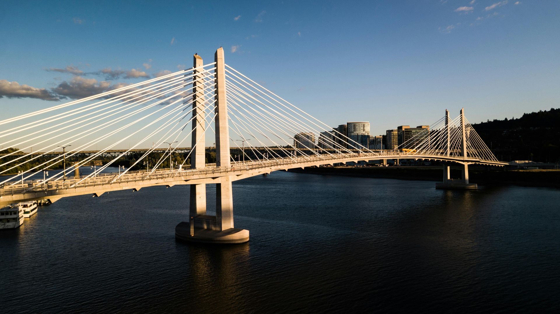 Cable-stayed bridge over water at sunset; city buildings visible in the distance.