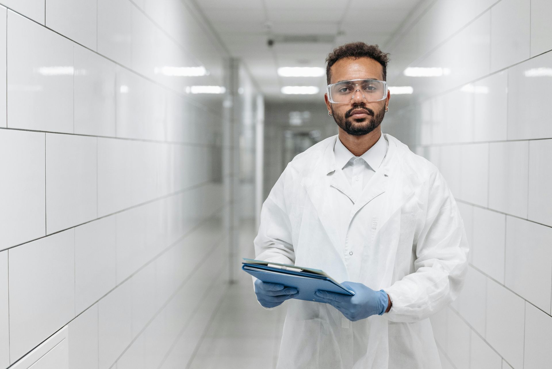 A man in a lab coat and goggles is holding a clipboard in a hallway.