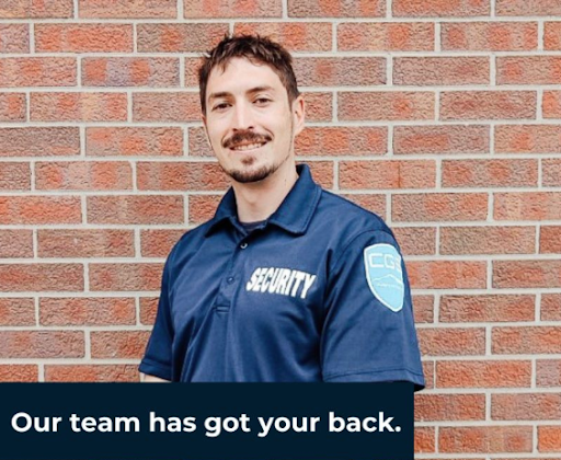 A man wearing a security shirt stands in front of a brick wall