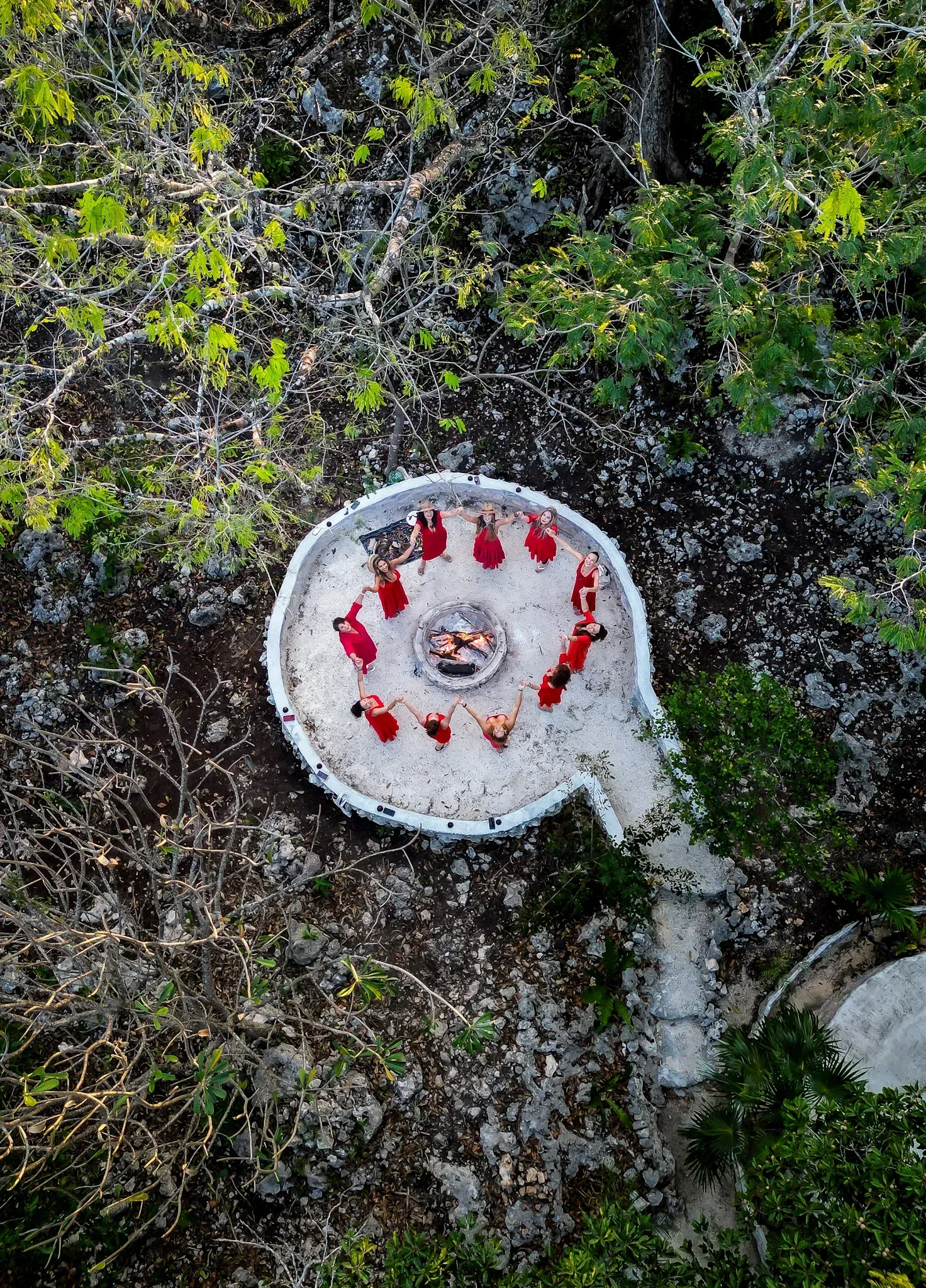 Vista aérea: Personas vestidas de rojo alrededor de un fuego en un círculo de piedras en una zona boscosa.