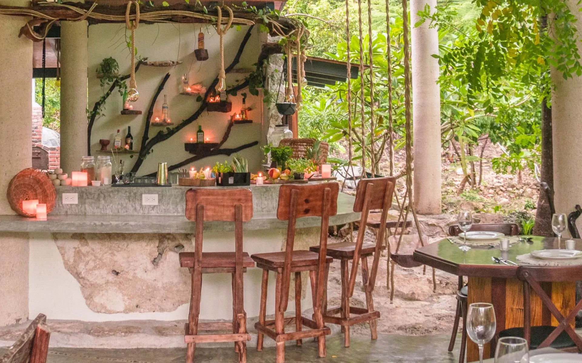 Bar al aire libre con taburetes y mesa de madera, rodeado de exuberante vegetación y velas.