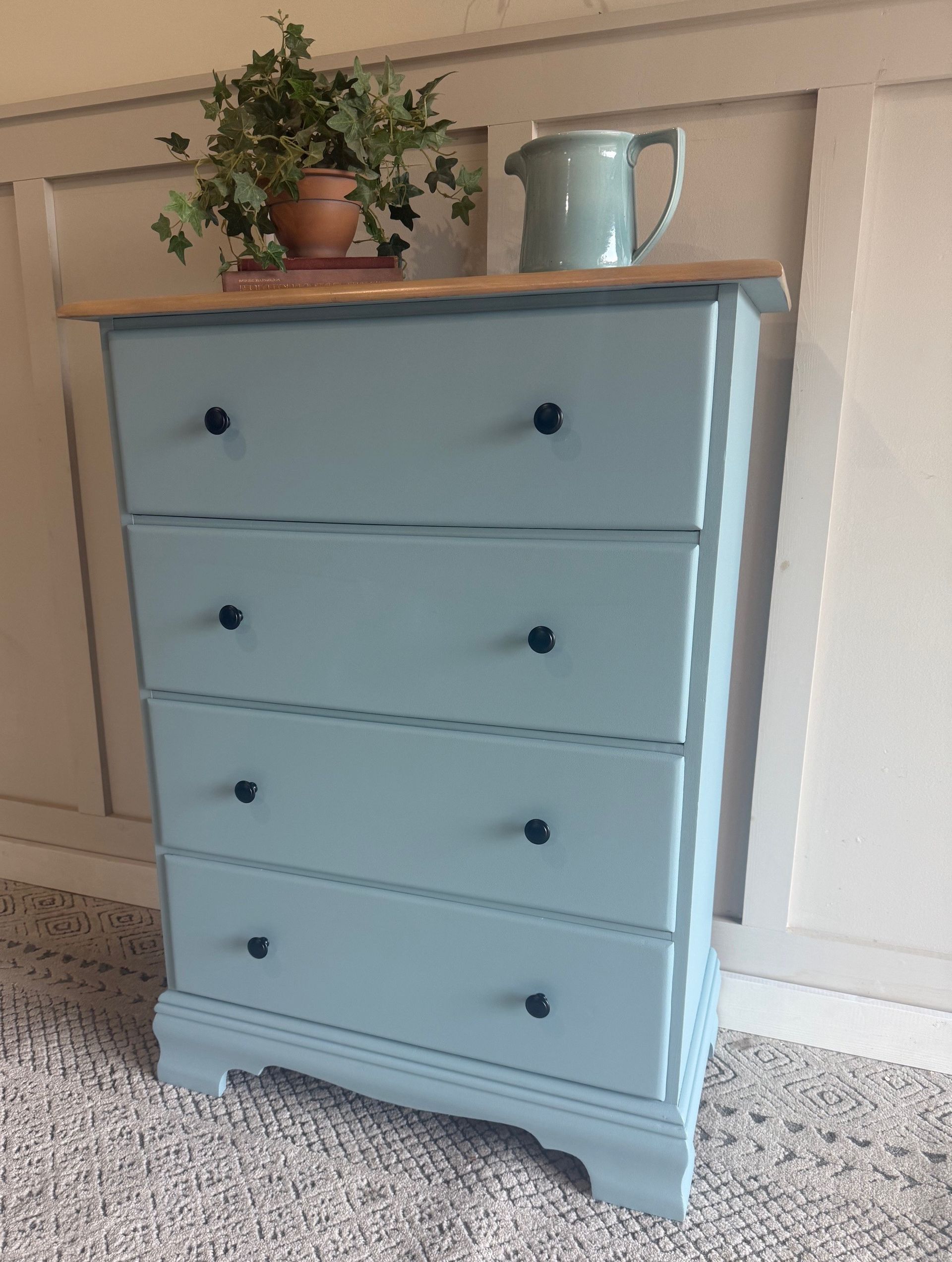 A blue painted dresser with four drawers, wooden top, and black knobs, with a plant and pitcher.