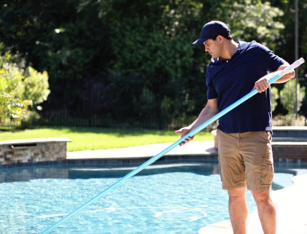Person using a pool skimmer to clean a backyard swimming pool on a sunny day