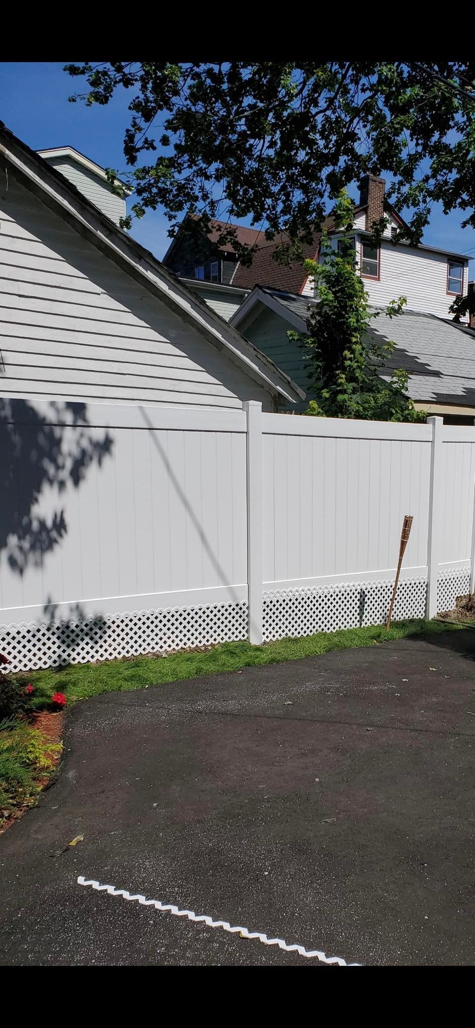 A white vinyl privacy fence with a lattice bottom section stands beside an asphalt driveway under a blue sky.