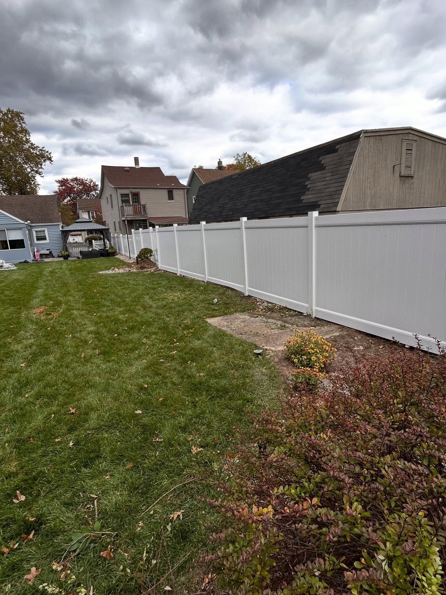 A grassy backyard scene features a tall white vinyl privacy fence running alongside a beige house with a dark roof.