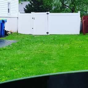 A white vinyl fence stands in a grassy backyard next to a house wall and a blue trash bin.