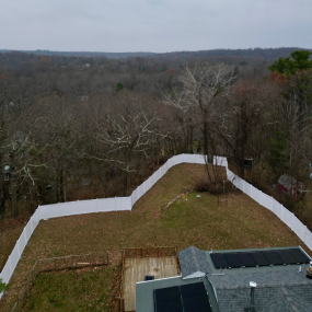 Aerial view of a house with solar panels on the roof and a large white fence enclosing a grass backyard facing the woods.