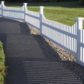 A white picket fence runs along a paved path bordered by a gravel strip next to a green lawn.
