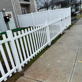 A white vinyl picket fence stands along a concrete sidewalk next to a house.