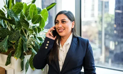 Woman in blue blazer, smiling, talking on a phone, near a window with plant.