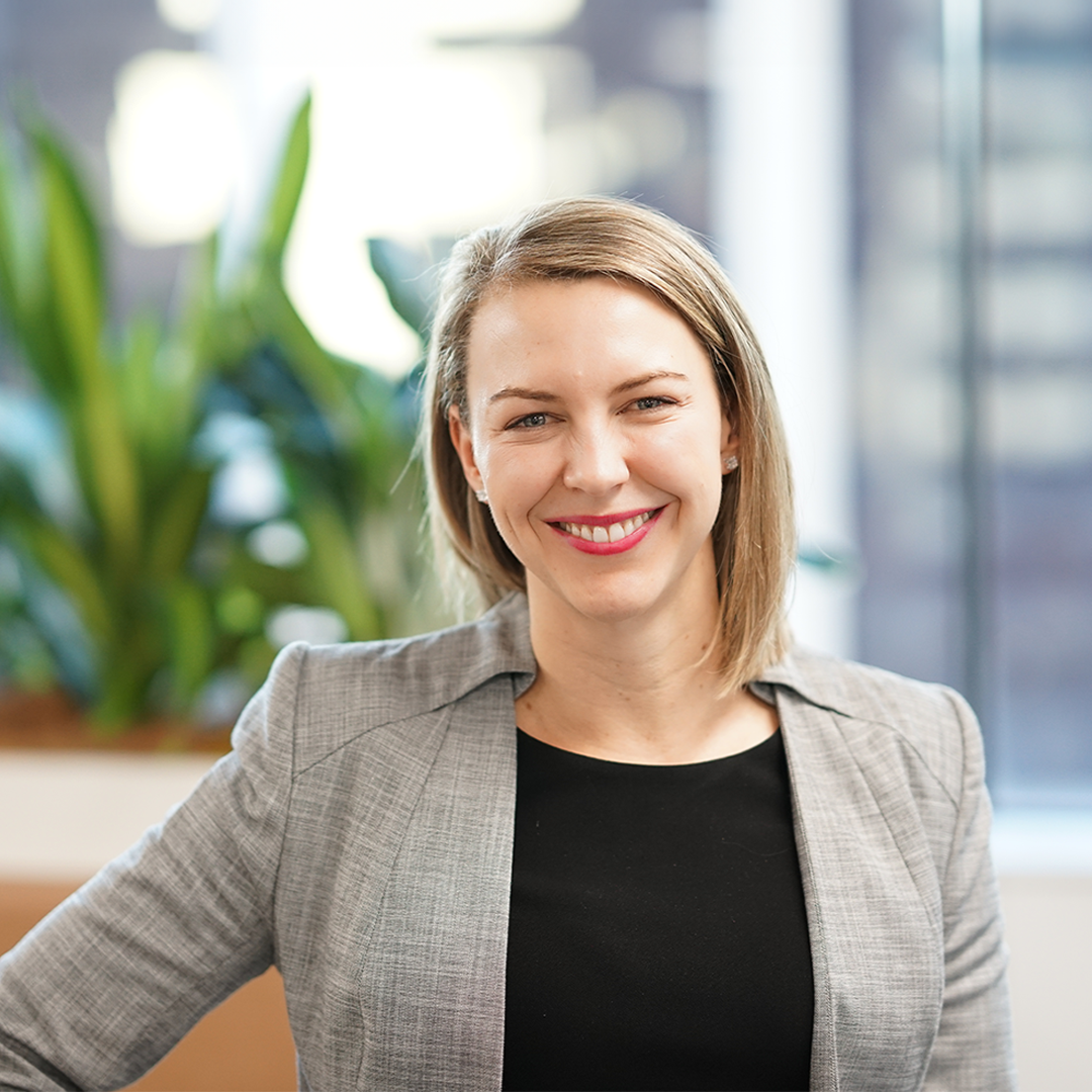 Woman smiling, wearing a gray blazer and black top. Indoor office setting with green plants.