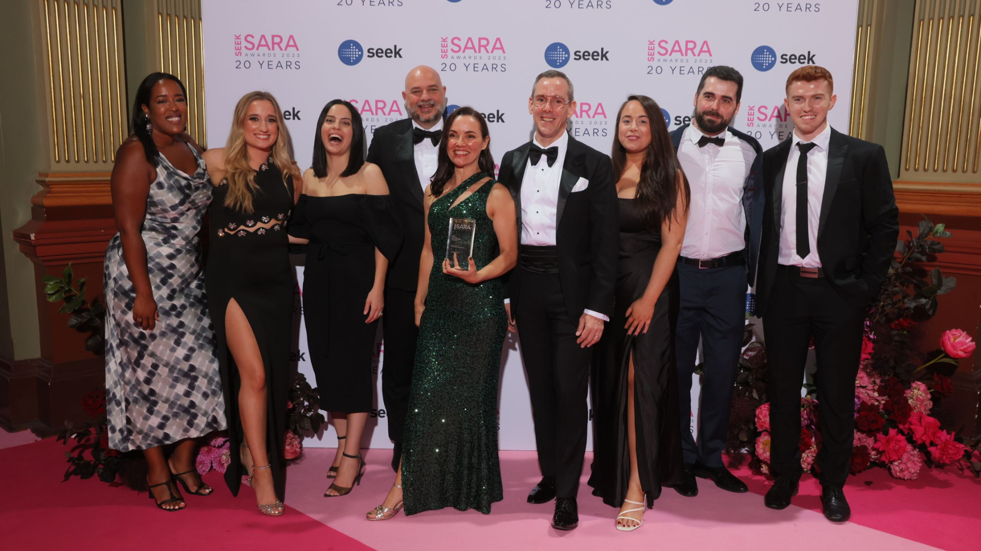 Group of people in formal attire pose for a photo at an awards event on a pink carpet.