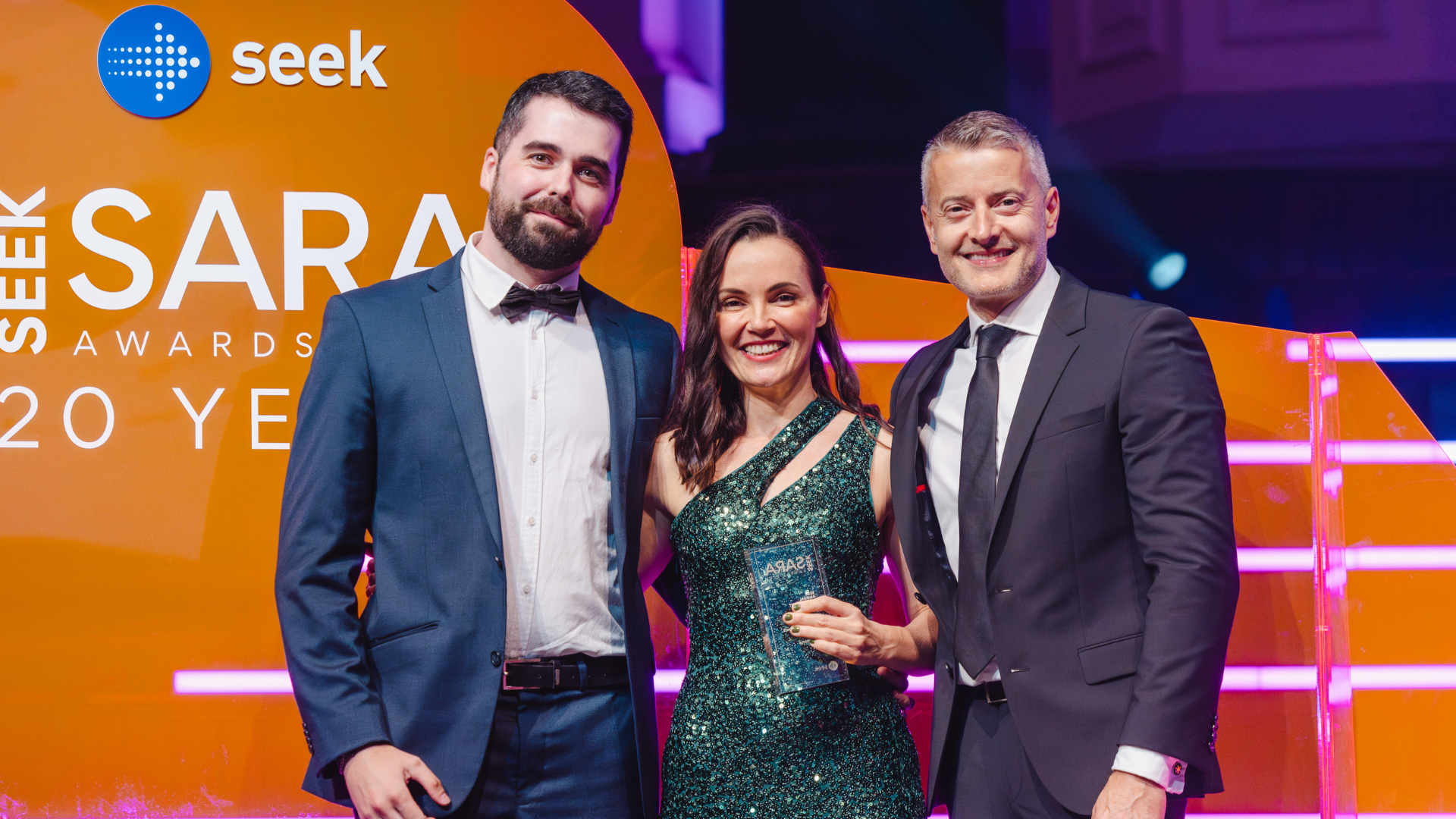 Three people on stage with award, woman in green dress holding it, 