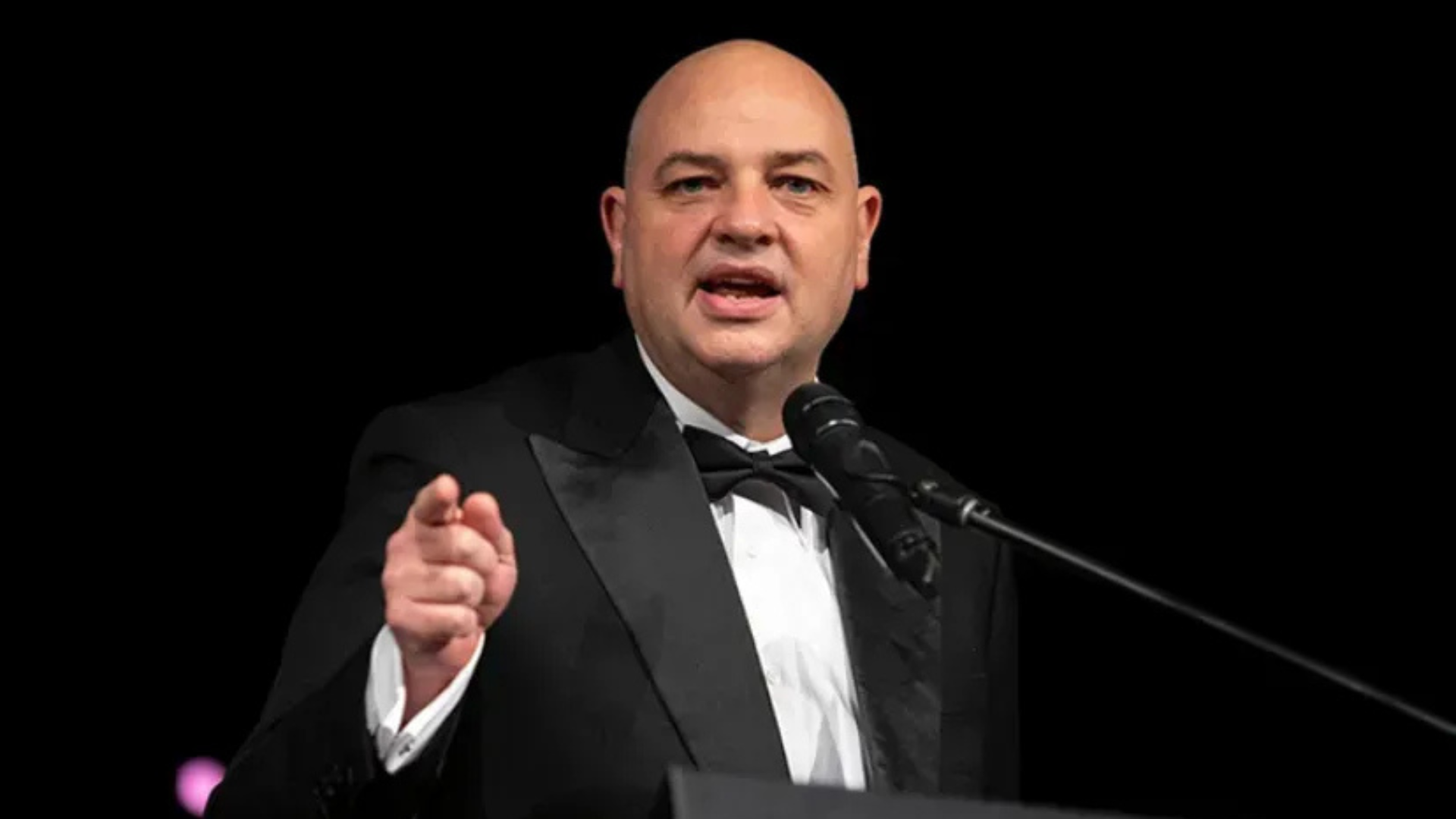 Man in a tuxedo speaking into a microphone, pointing, black background.