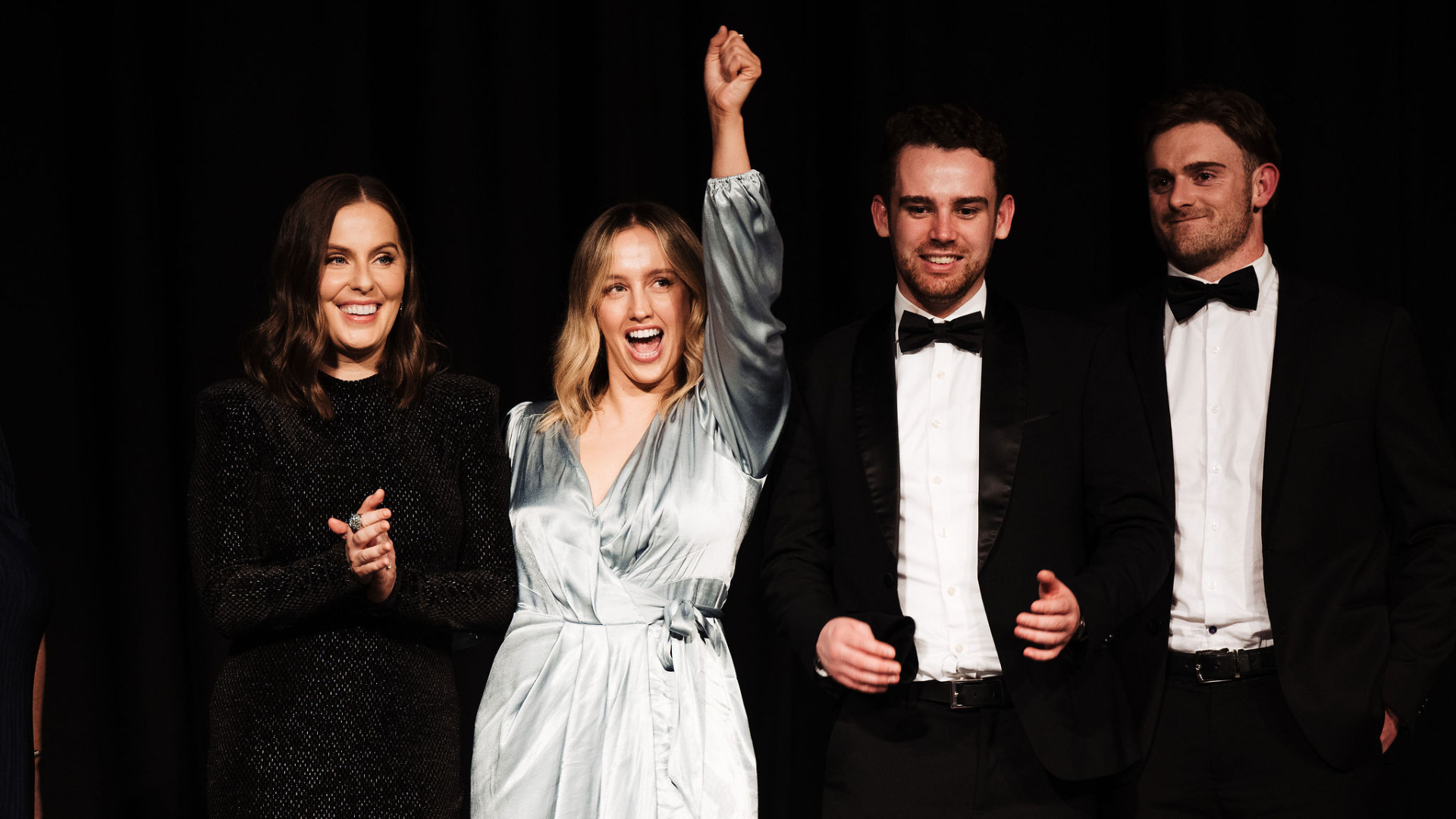 Four people on a stage. One raises fist, smiling. Others clap and smile in formal wear. Dark backdrop.